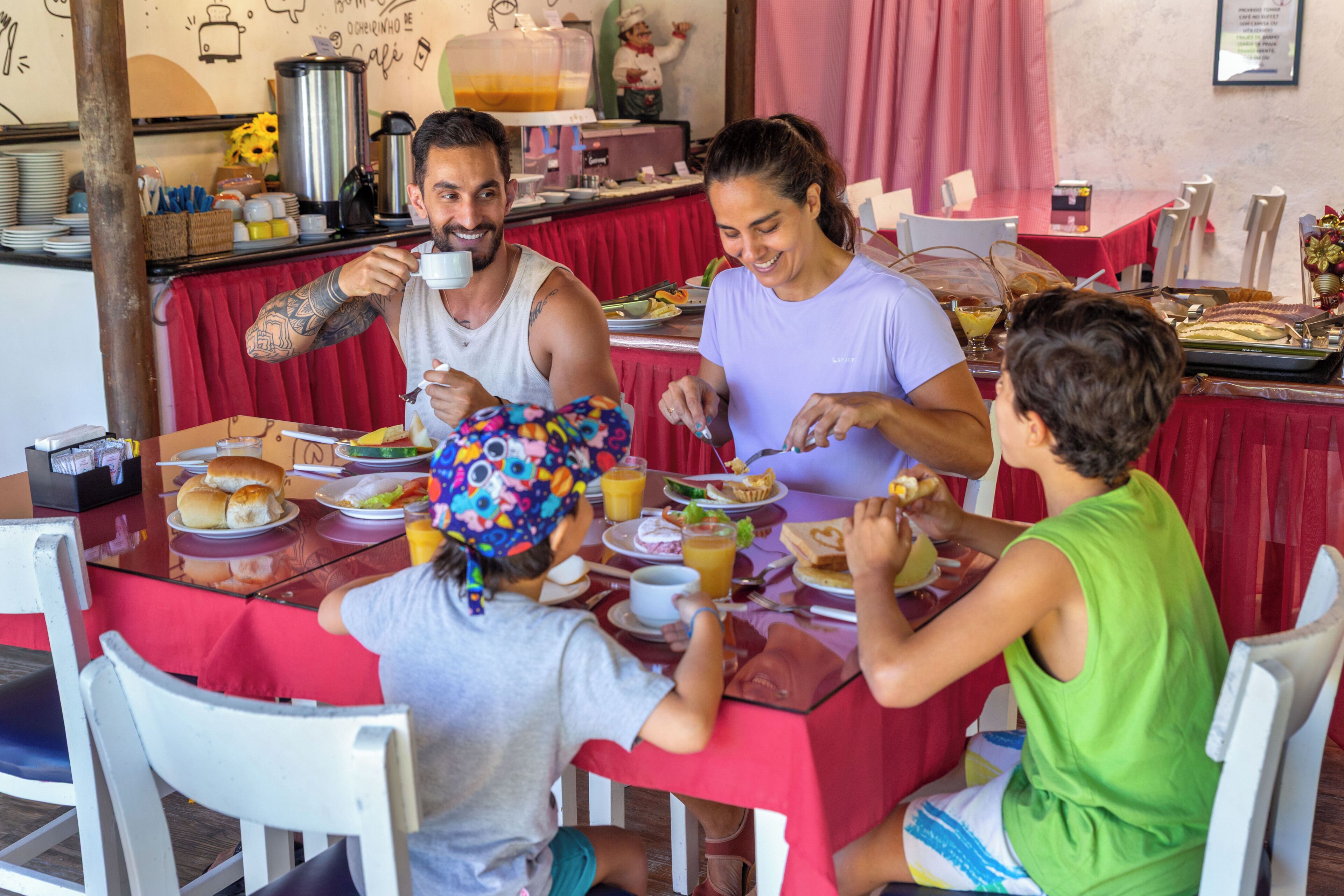 Eine Familie sitzt an einem Tisch mit rotem Tischtuch, es gibt verschiedene Gerichte und im Hintergrund steht eine Kaffeemaschine.