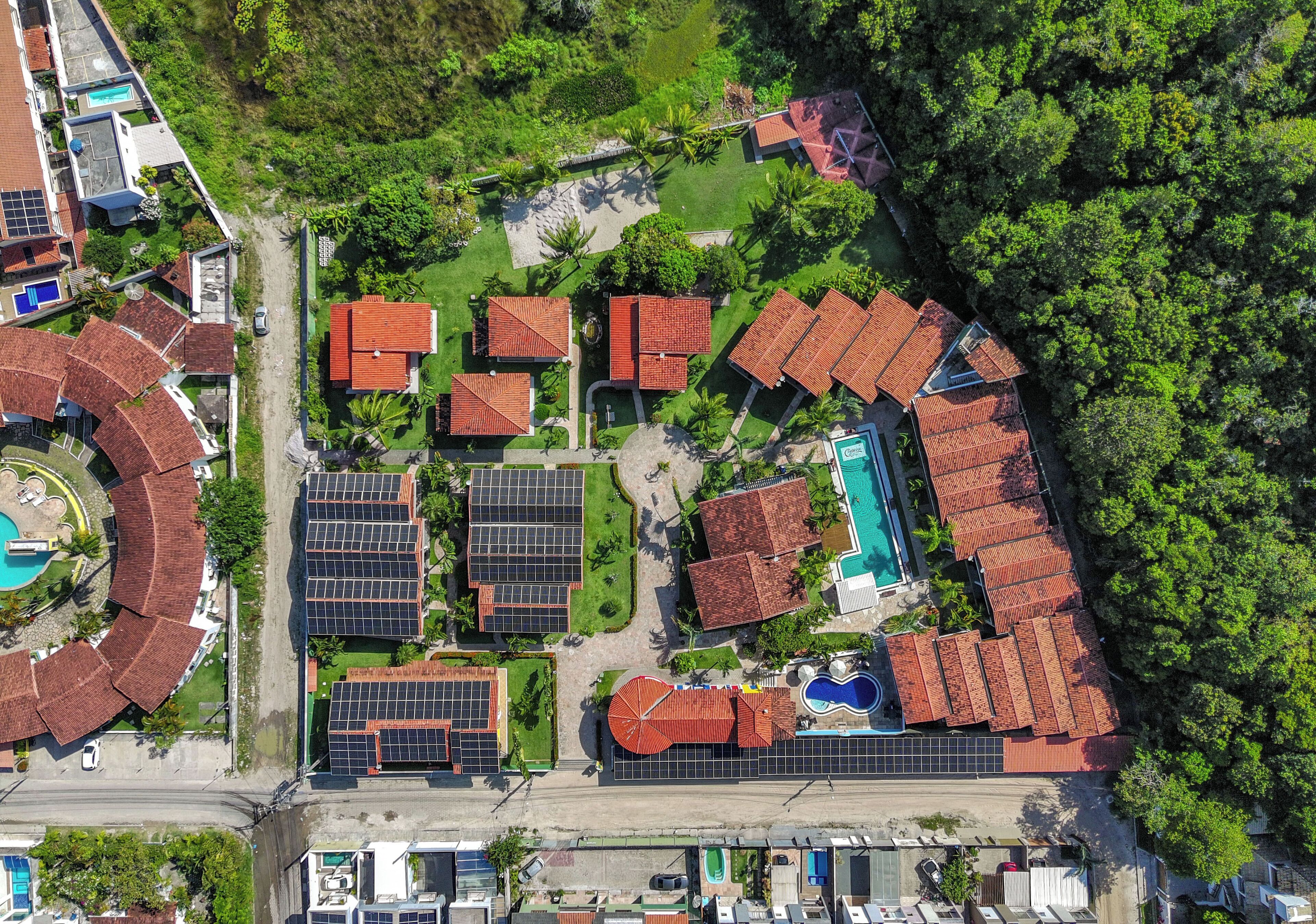 An aerial view of a residential area with multiple houses, solar panels, a swimming pool, and lush greenery.