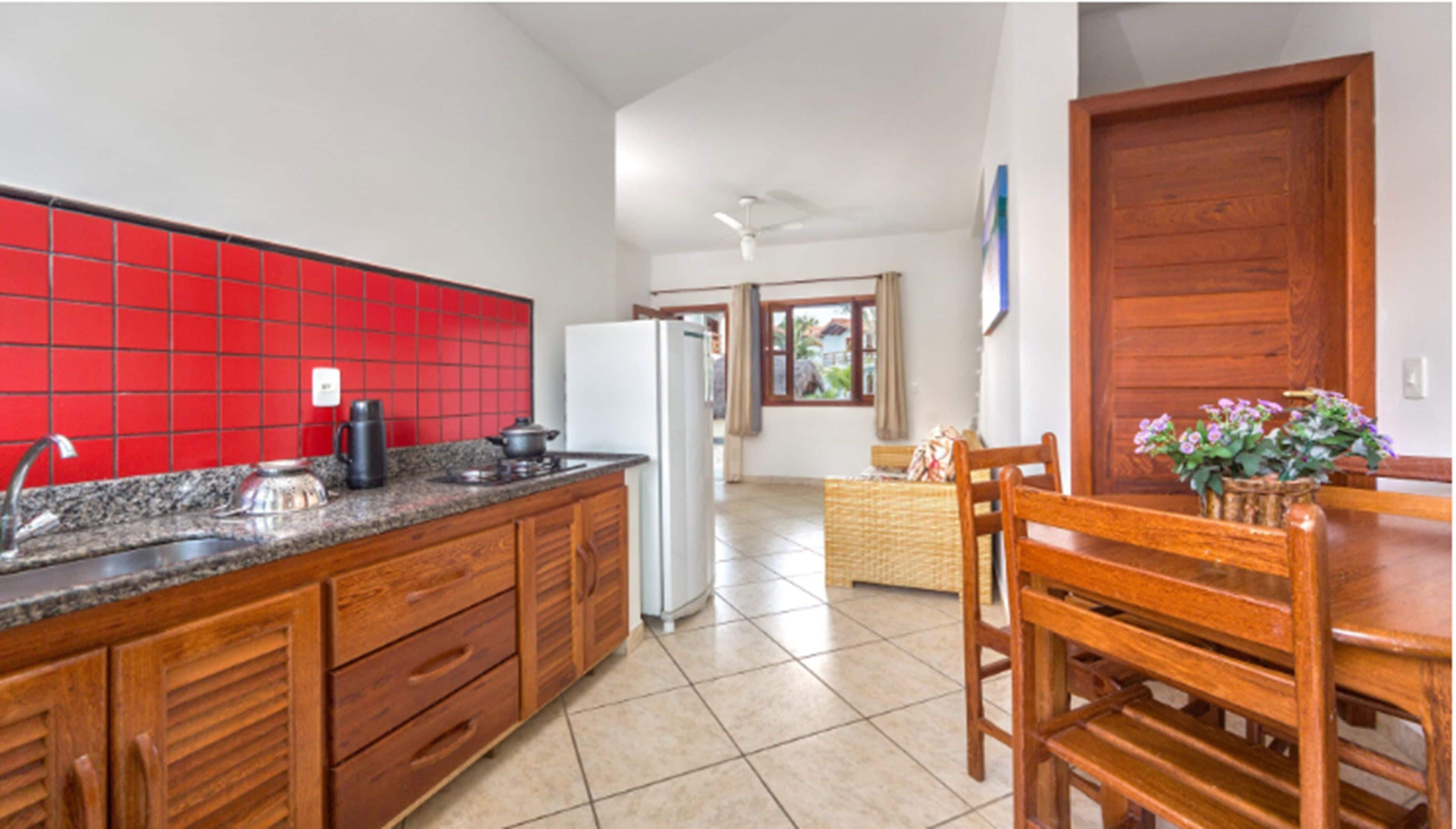 A kitchen with red tiles, a refrigerator, a stove, and a sink. There is a dining table with chairs and a window with curtains.