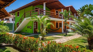 Une maison à deux étages avec une façade verte et orange, un balcon en bois et un jardin bien entretenu agrémenté de fleurs rouges.