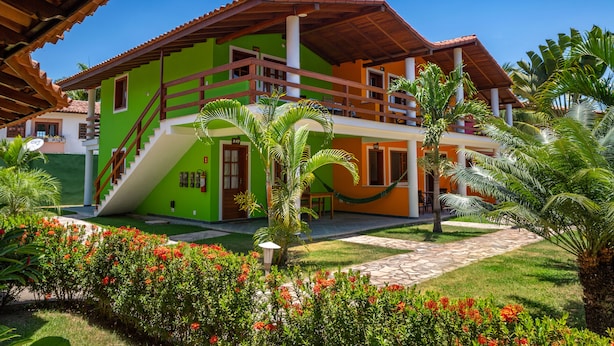 A two-story house with a green and orange facade, a wooden balcony, and a well-maintained garden with red flowers. - Capoeira Village (Porto Seguro)