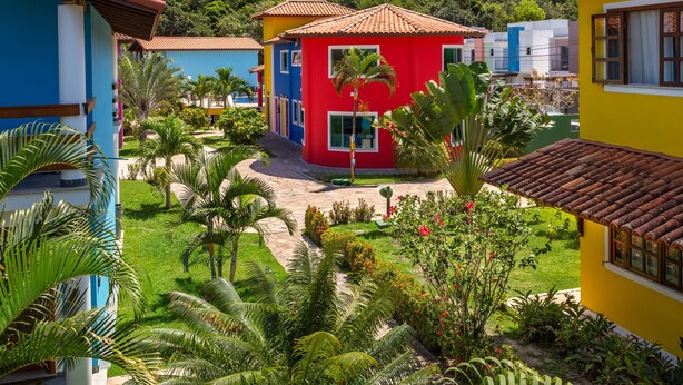 A colorful building with a red, blue, and yellow facade, surrounded by lush greenery and a paved pathway. - Capoeira Village (Porto Seguro)