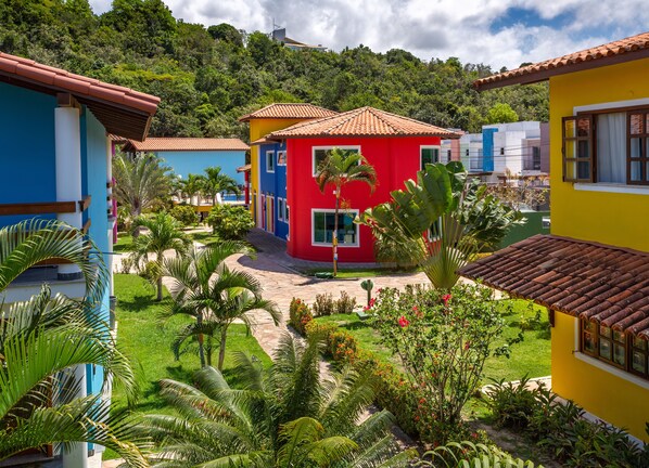 A colorful building with a red, blue, and yellow facade, surrounded by lush greenery and a paved pathway. - Capoeira Village (Porto Seguro)