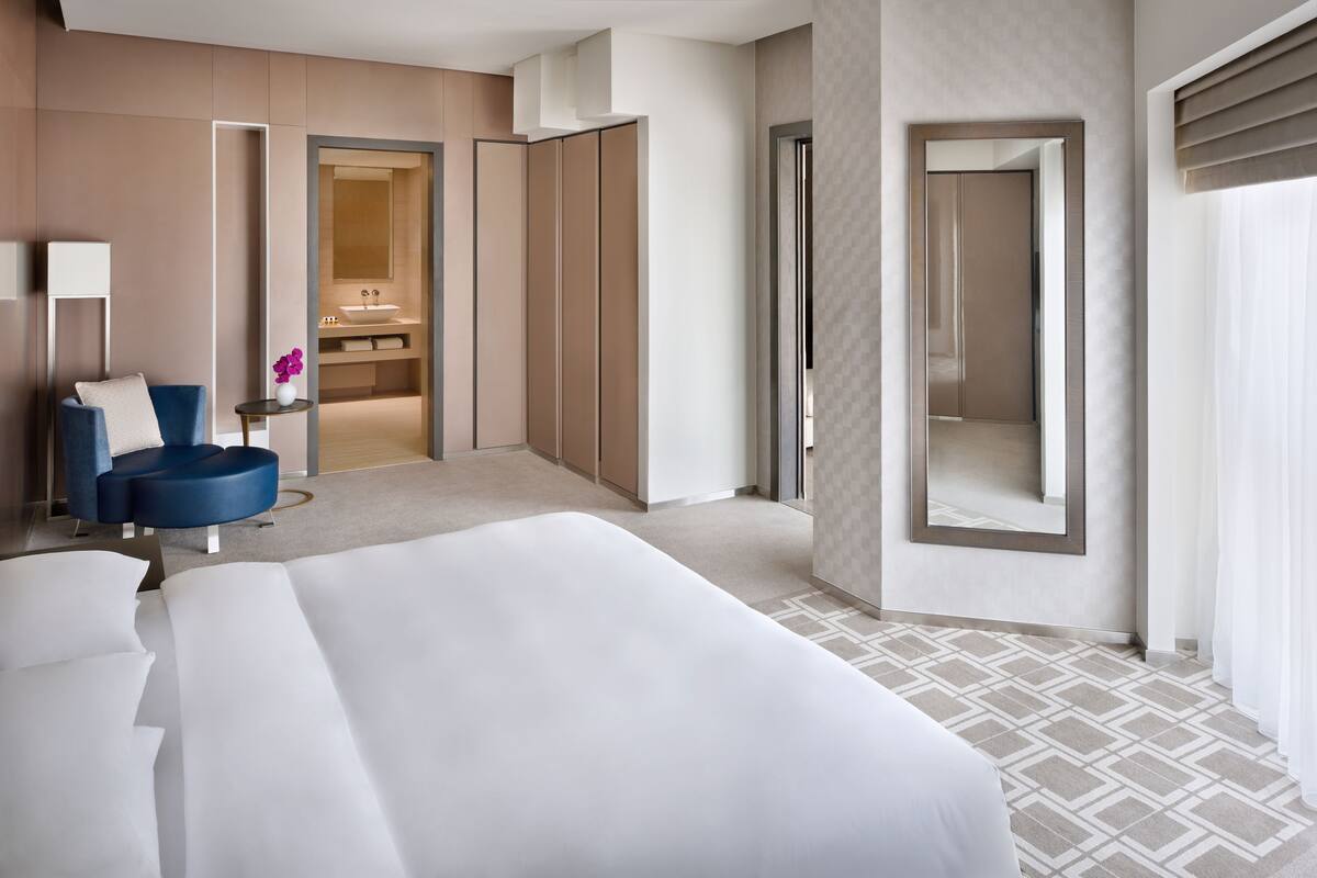 Habitación de hotel con una cama grande, una silla azul, una mesita con un florero y un baño con espejo y lavamanos.