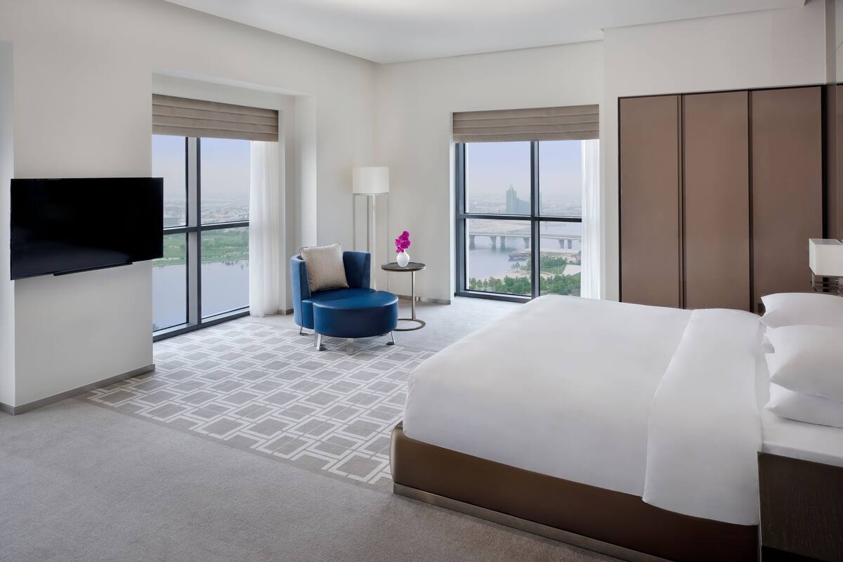 Habitación de hotel con una cama grande, un televisor montado en la pared, una silla, una mesita con lámpara y ventanas amplias con vistas a la ciudad.