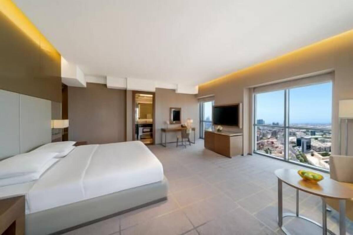 Habitación de hotel con cama, escritorio, televisión y ventana grande con vista a la ciudad.