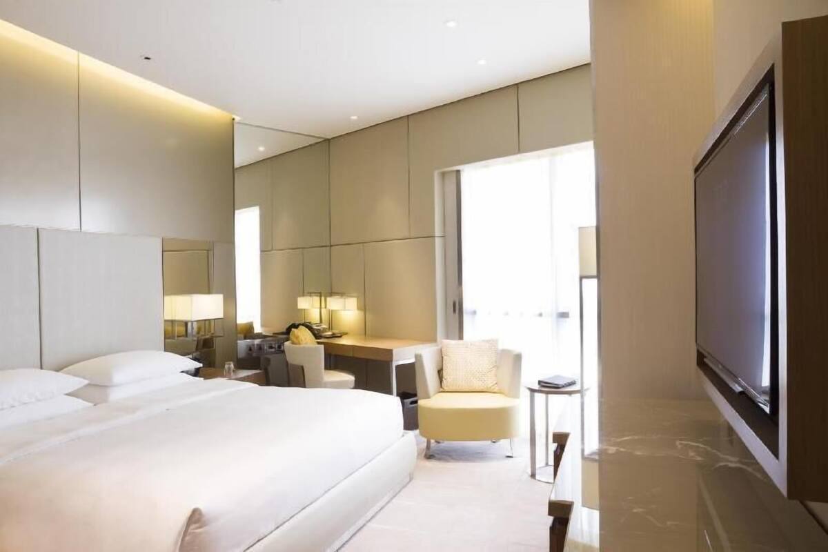 Una habitación de hotel moderna con una cama grande, un escritorio, una silla, un televisor y un ventanal amplio.