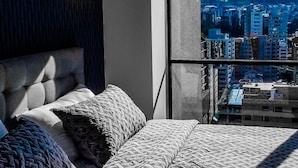 Quarto com cama grande, luminária pendente e vista para a cidade pela janela.