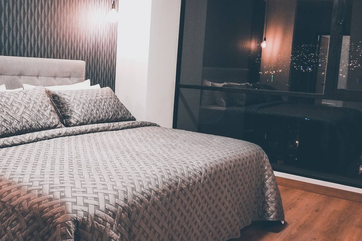 A neatly made bed with a patterned bedspread and pillows, a tufted headboard, and a view of a cityscape through a large window.
