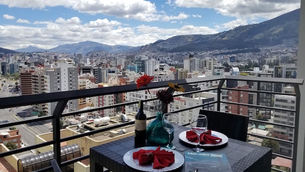 Varanda com mesa posta para dois, com vista para paisagem urbana e montanhas ao fundo.