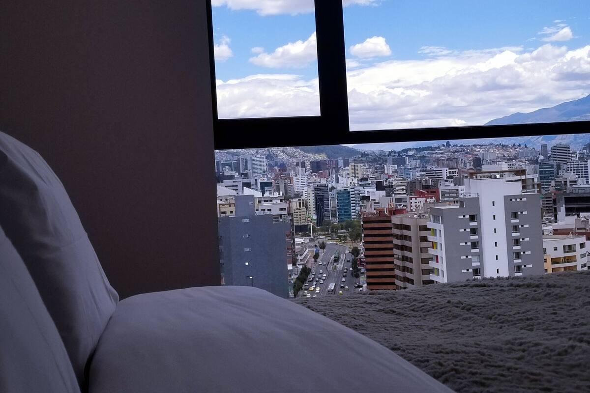 A hotel room with a large window overlooking a cityscape and mountains.