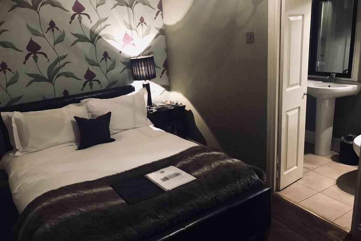 A neatly made bed with a fluffy blanket, a bedside lamp, and a floral-patterned wall. A door leads to a bathroom with a sink and mirror.