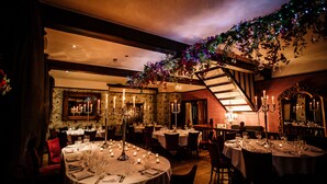 Un restaurante tenuemente iluminado con mesas redondas dispuestas para comer, centros de mesa con velas y una escalera adornada con una guirnalda decorativa.