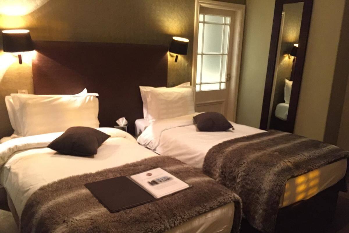 A hotel room with two beds, each with white linens and dark pillows. There are two wall-mounted lamps on either side of the beds. A window with frosted glass is visible in the background.