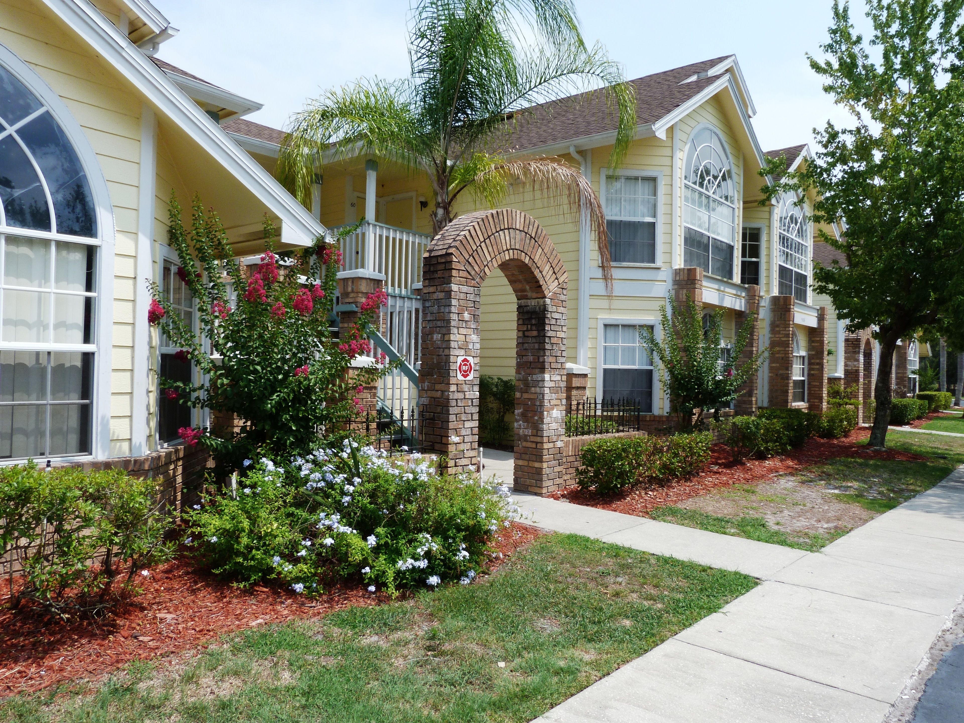 A residential building with a brick archway entrance, a covered porch, and a well-maintained garden with flowering plants.