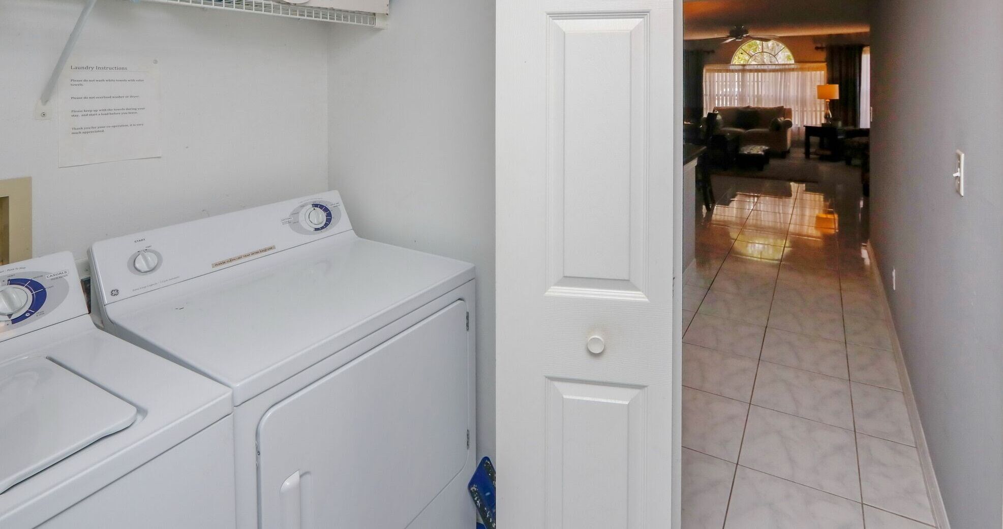 Un espace buanderie avec un lave-linge et un sèche-linge blancs, une porte donnant sur un salon équipé d’un canapé et d’une lampe.