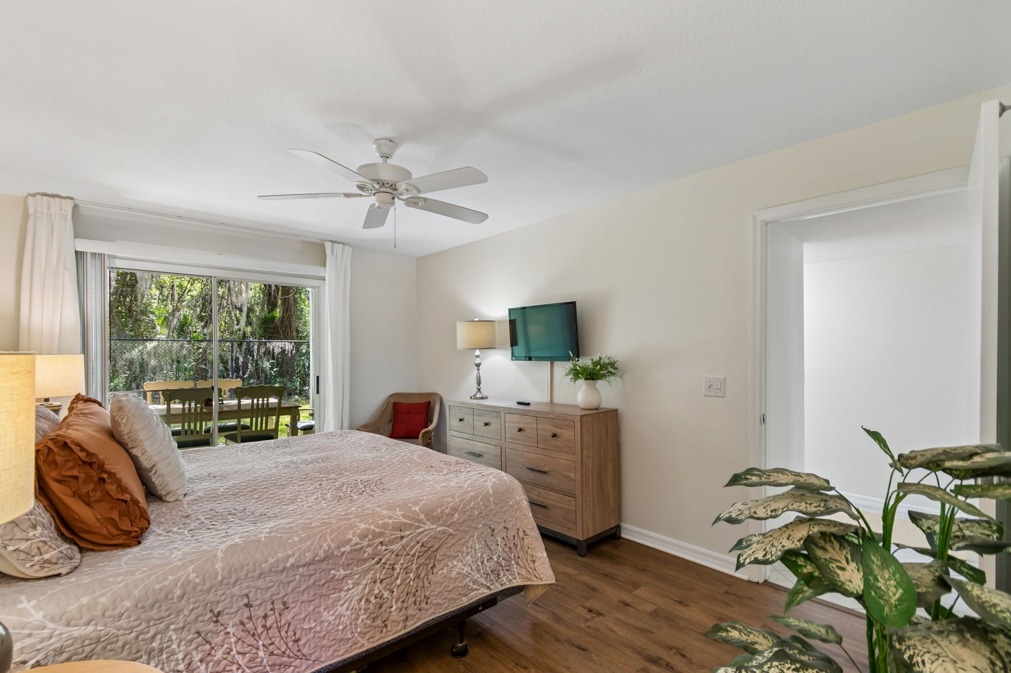 A bedroom with a bed, a ceiling fan, a TV, a dresser, a chair, and a window with a view of greenery.