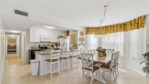 Een moderne keuken met witte kasten, een eethoek met een houten tafel en witte stoelen, en een gordijn met een geel patroon.