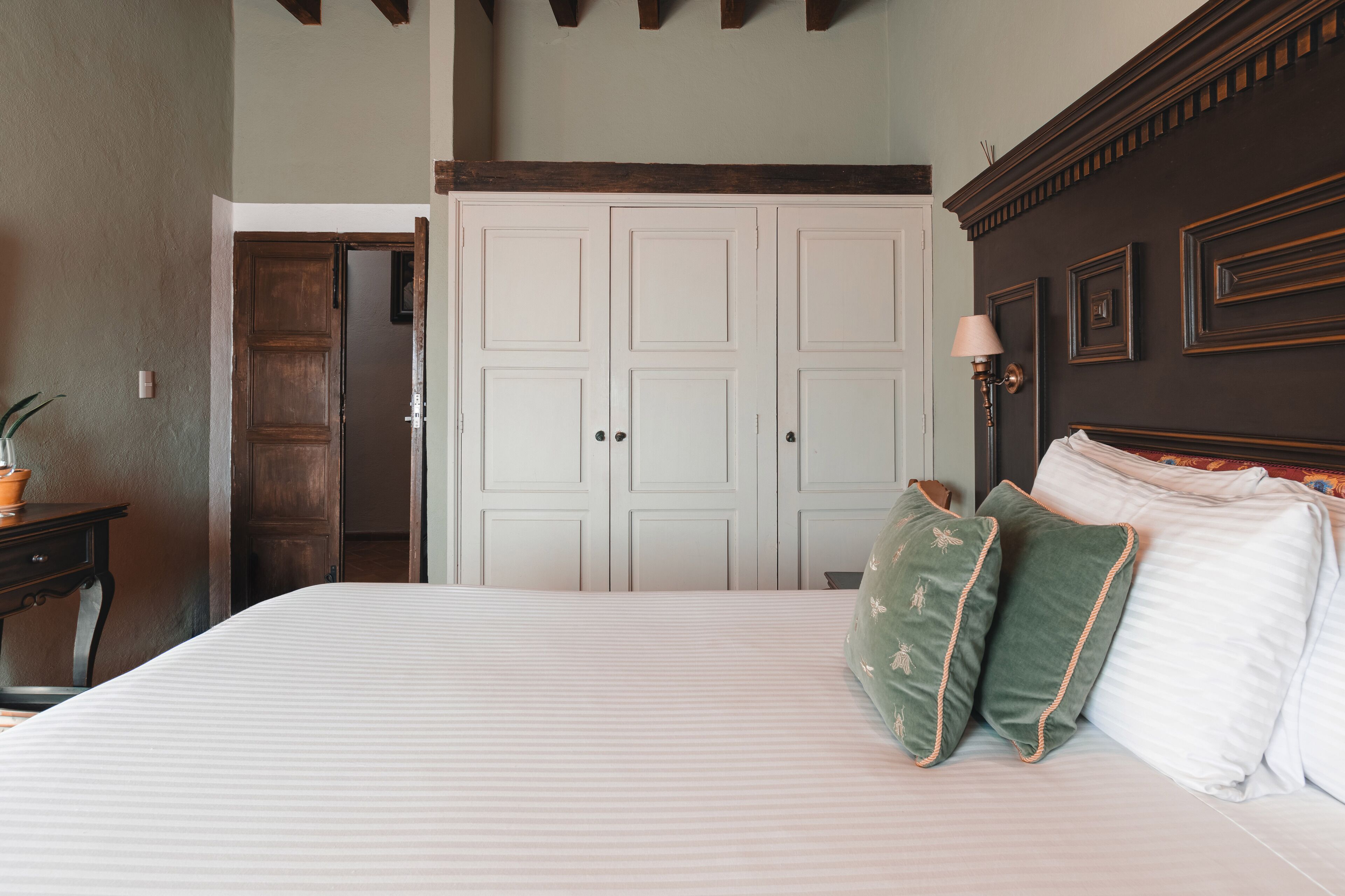 Un dormitorio con una cama grande, un armario de madera, una mesita de noche con una lámpara y una planta en maceta.