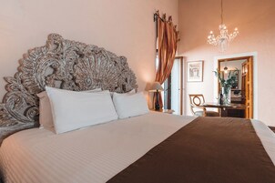 A bedroom with a large ornate headboard, a bed with white and brown bedding, a chandelier, and a view of a dining area.