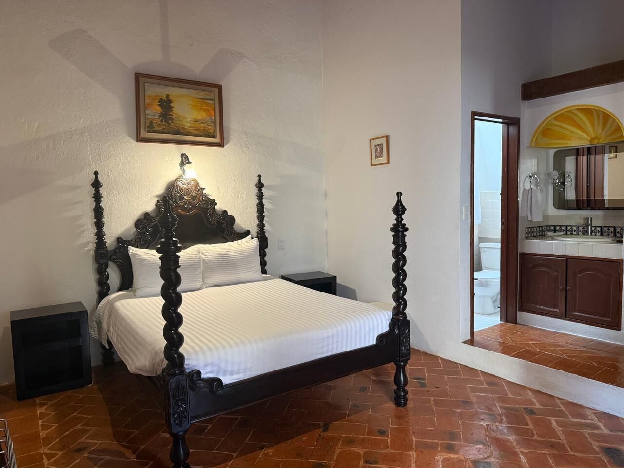 A bedroom with a four-poster bed, a nightstand, a painting on the wall, and a view into the bathroom.