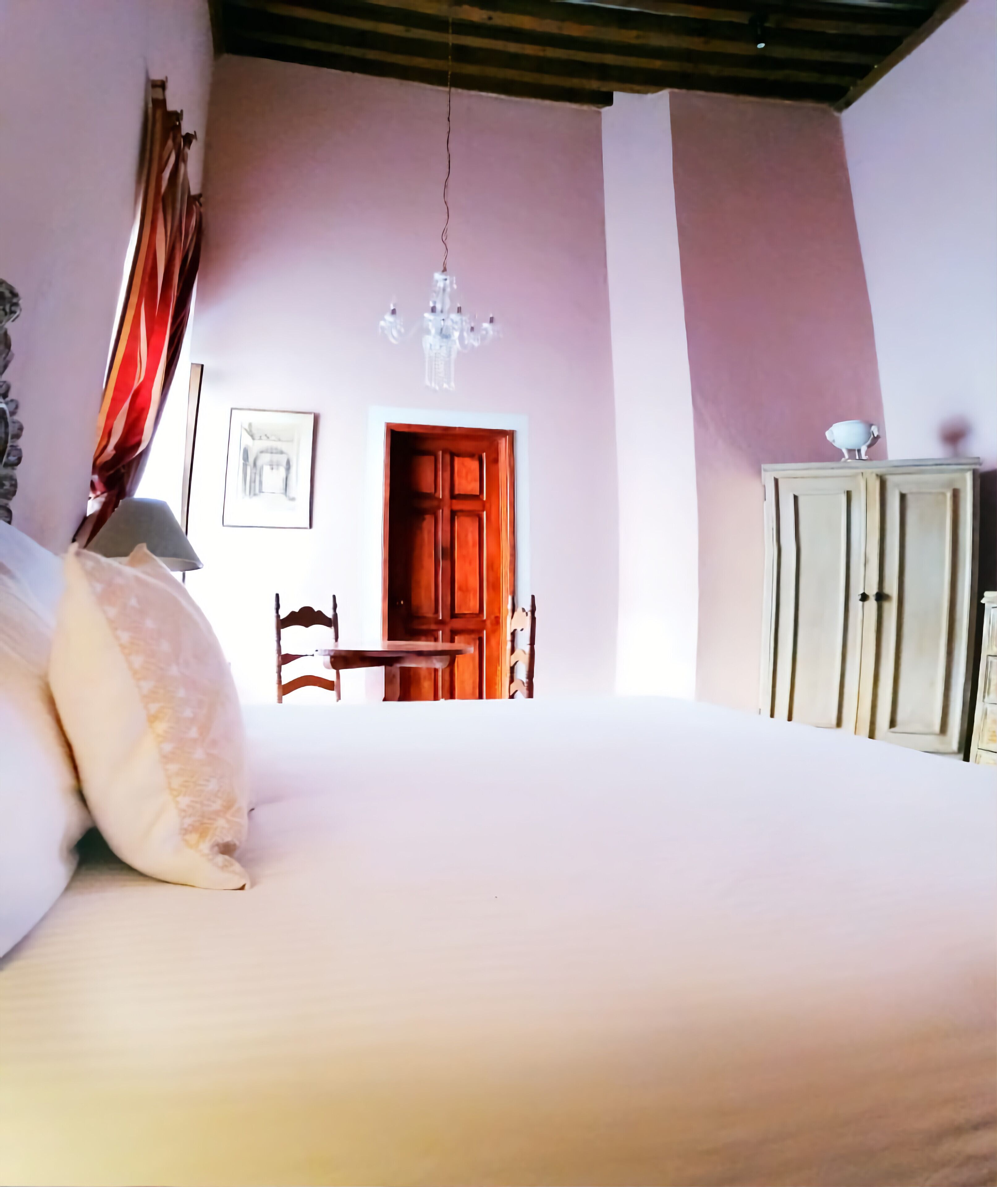 A bedroom with a bed, a wooden chair, a wooden wardrobe, a door, and a chandelier.