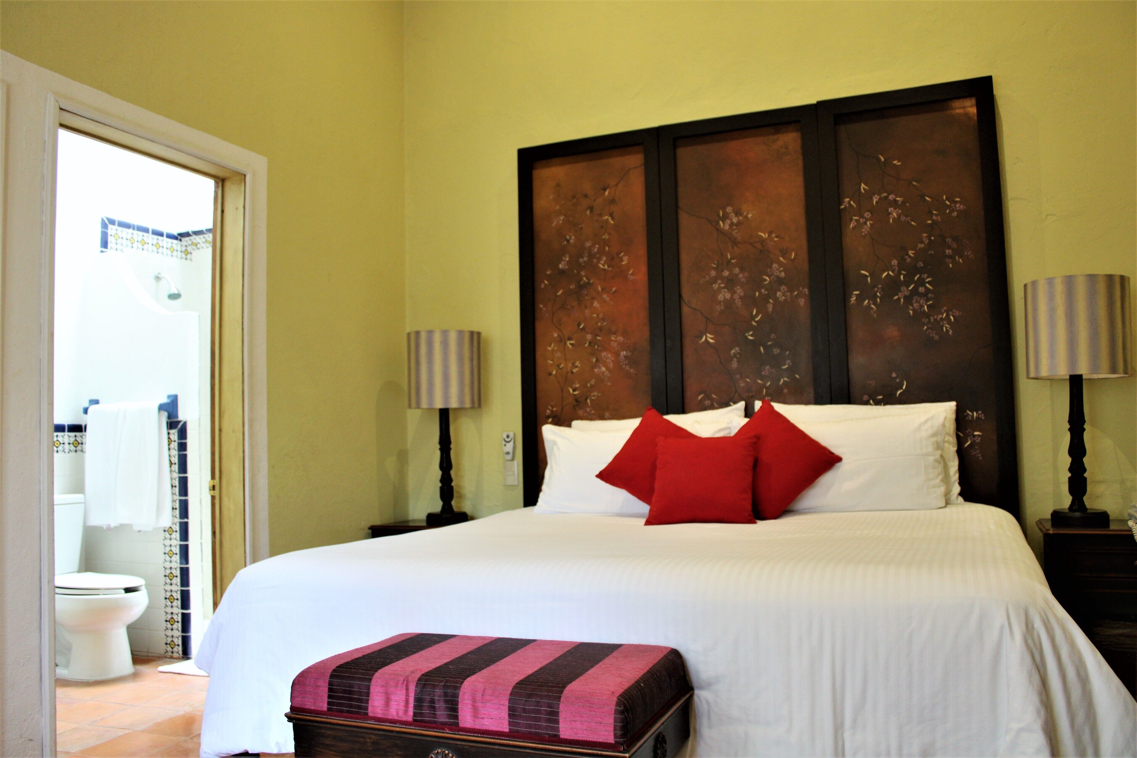 A neatly made bed with white linens and a red pillow, a wooden headboard, two bedside lamps, and a small wooden chest at the foot of the bed.