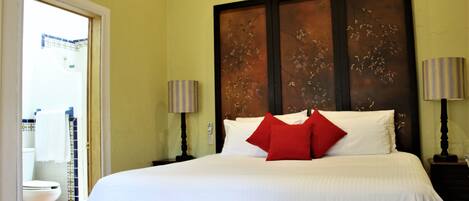 A neatly made bed with white linens and a red pillow, a wooden headboard, two bedside lamps, and a small wooden chest at the foot of the bed.