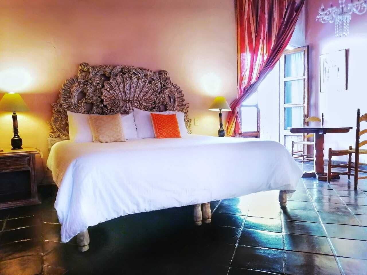 A bed with a white bedspread and multiple pillows, a wooden headboard, two bedside lamps, and a window with curtains.