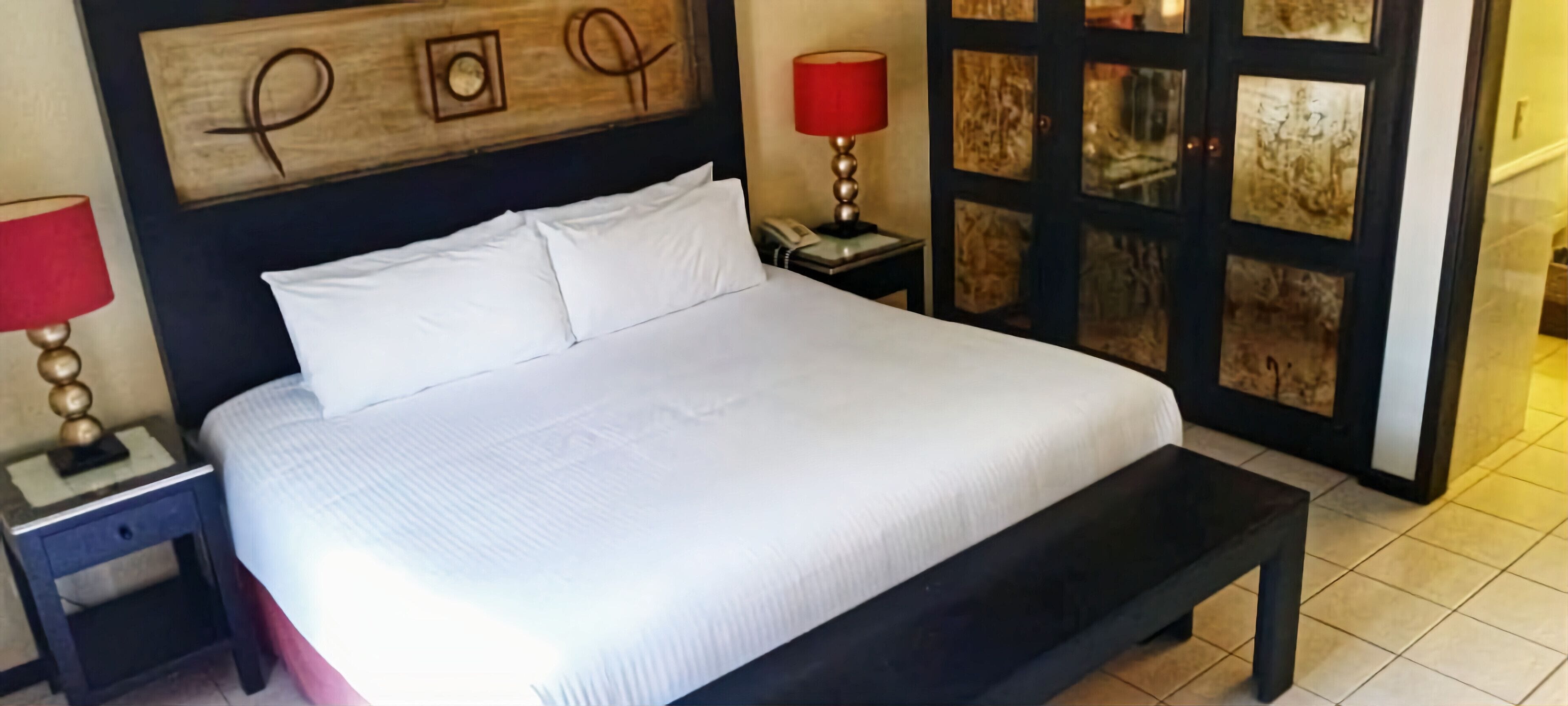 A neatly made bed with white linens, a wooden headboard with a decorative plaque, a bedside table with a lamp, and a nightstand with a telephone.