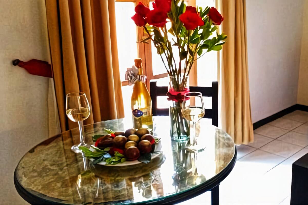 Habitación de hotel con cama, una mesa con un florero, una botella de vino y un plato de fruta.