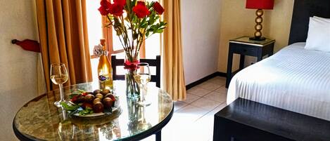 Quarto de hotel com cama, uma mesa com um vaso de flores, uma garrafa de vinho e uma travessa de fruta.