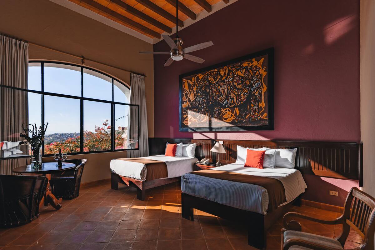 Habitación de hotel con dos camas, una mesa de comedor, sillas, un ventilador de techo y un ventanal con vistas.