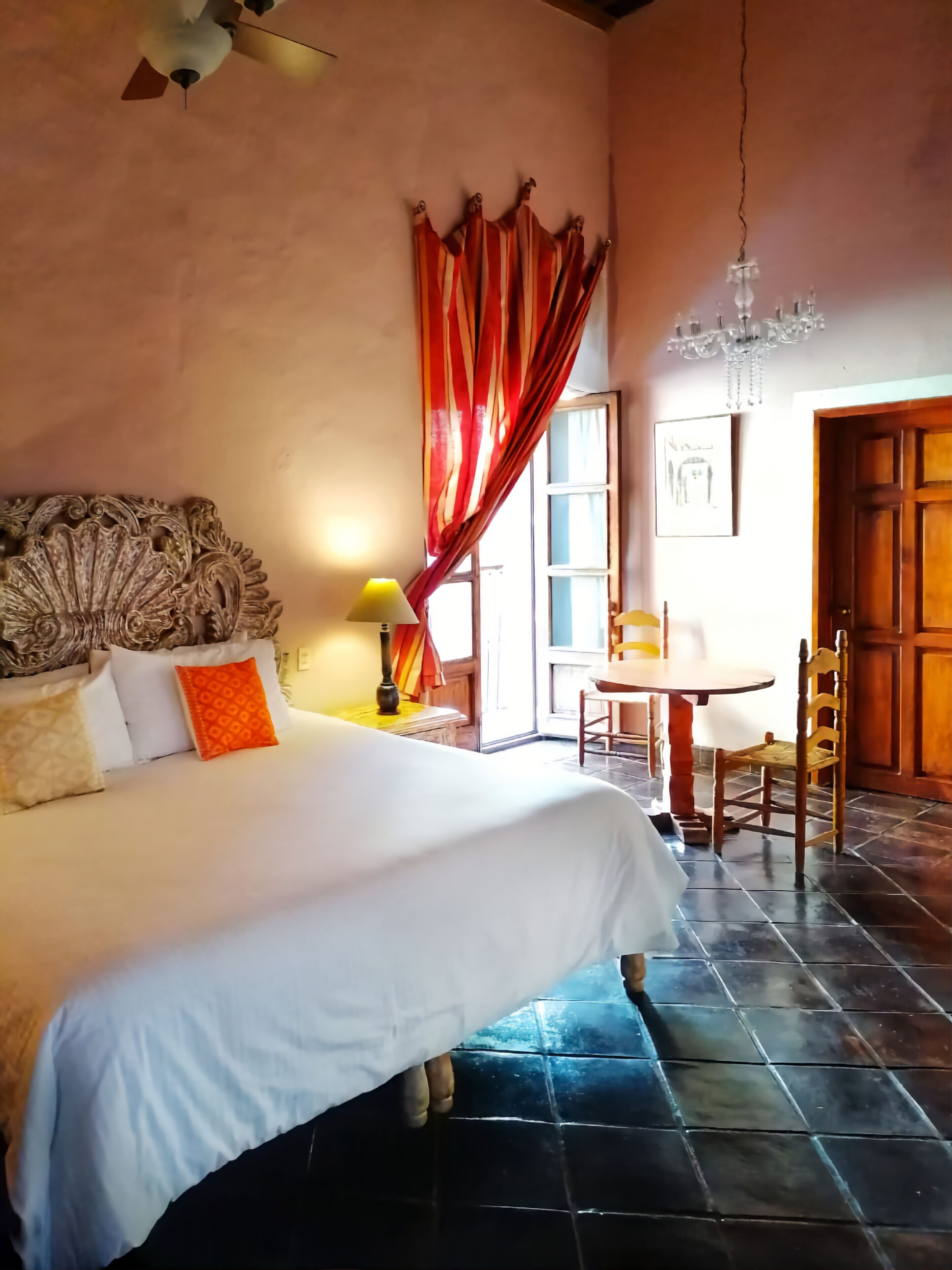 Un dormitorio con una cama grande, cabecera de madera, una lámpara de araña, una mesa con sillas y vistas al exterior a través de una ventana con cortinas rojas.