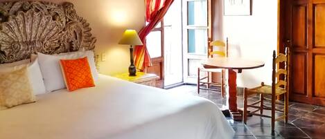 A bedroom with a large bed, a wooden headboard, a chandelier, a table with chairs, and a view of the outside through a window with red curtains.