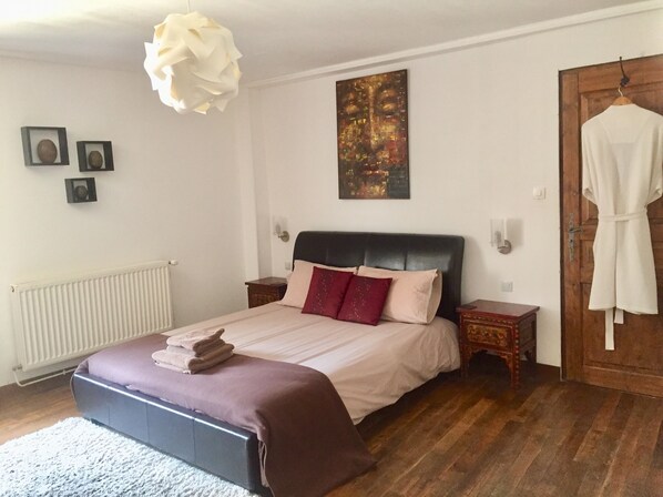 A bedroom with a bed, a radiator, a wooden floor, a white robe hanging on a door, and a painting on the wall.