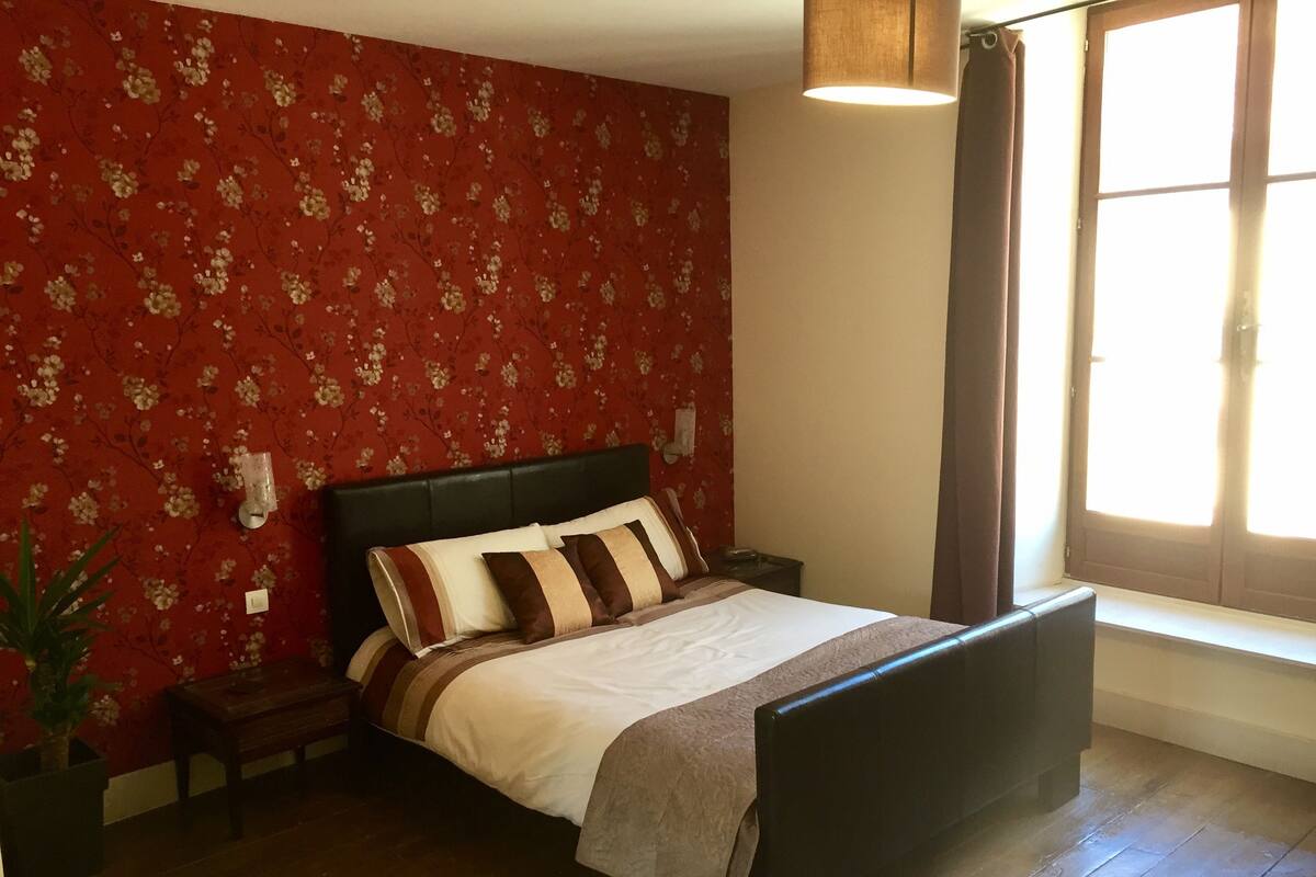 Ein Schlafzimmer mit blumigem Wandpapier, einem Bett mit gestreiften Kissen, einem Nachttisch, einer Stehlampe und einem Fenster mit Vorhängen.