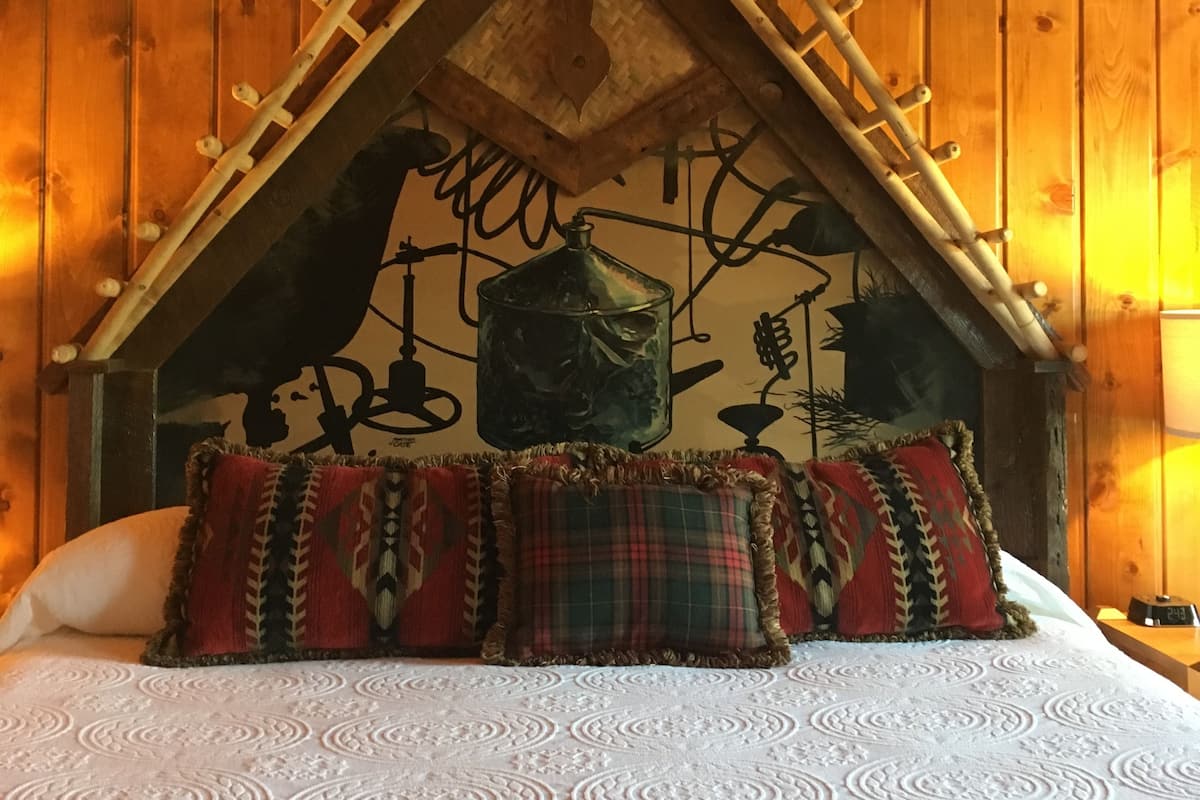 Une pièce aménagée comme un chalet en bois, avec un plafond en pente, un lit recouvert d’une literie à motifs et une fresque murale représentant une scène champêtre.