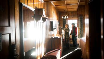 A cabin interior with wooden walls, a bench, a small table, and a window with curtains.