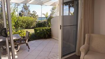 A room with a sliding glass door leading to a patio with a table and chair, a beige sofa, and a view of greenery and a body of water.