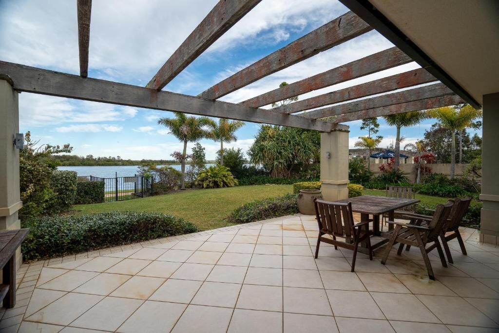 A covered patio with a table and chairs, overlooking a body of water and a well-maintained lawn.