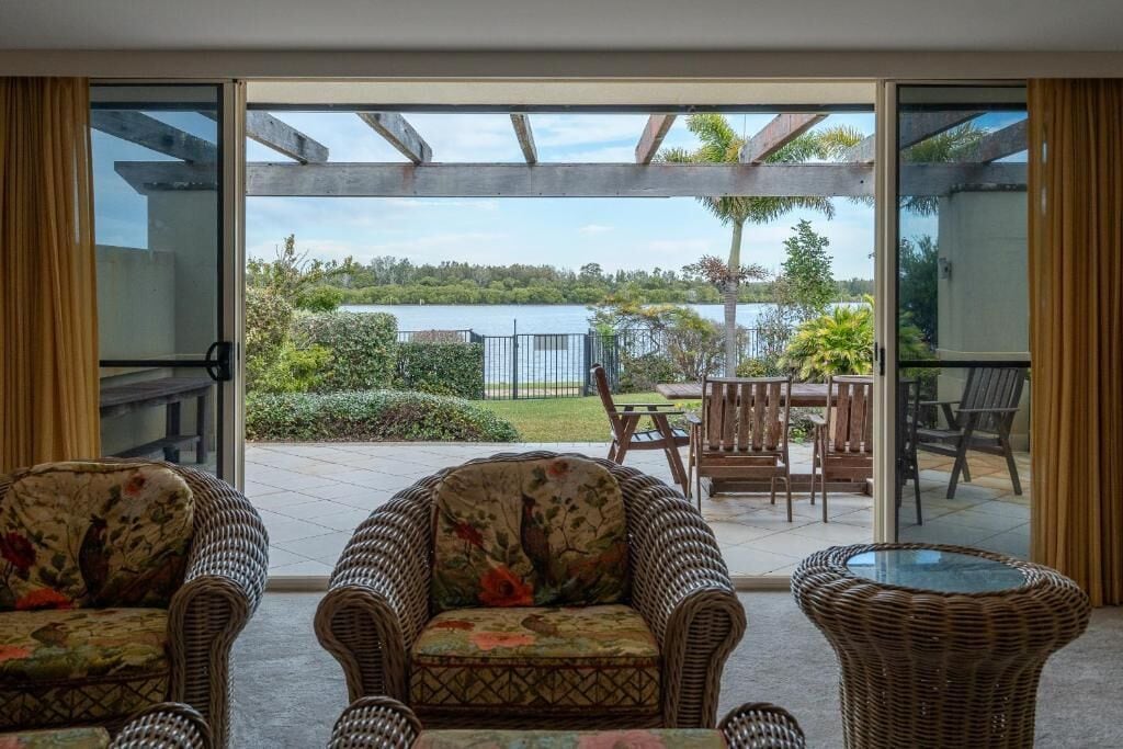 A room with wicker furniture, a glass door leading to an outdoor patio with a view of a lake and greenery.