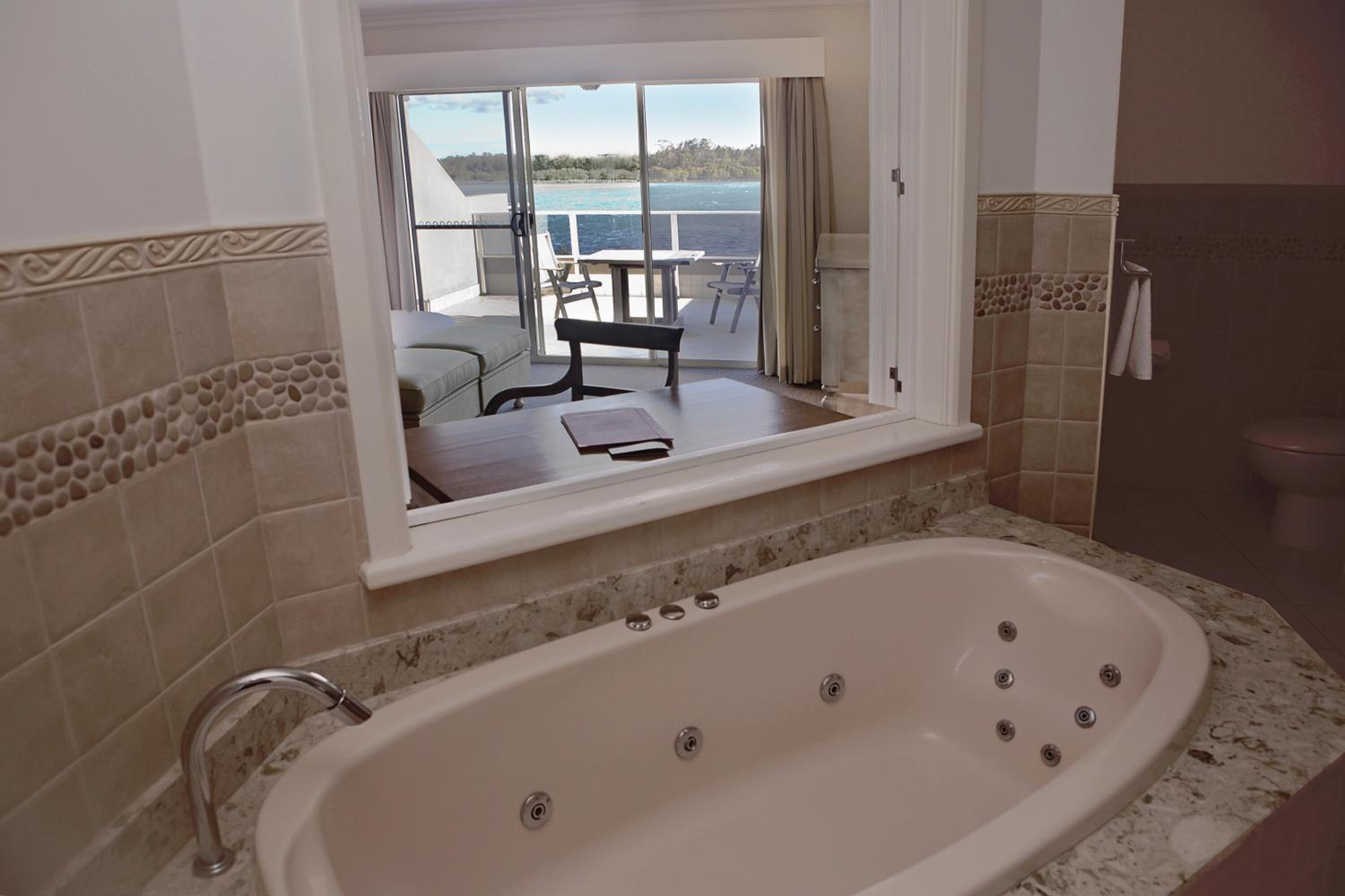 A bathroom with a large bathtub, a window overlooking a deck with outdoor furniture, and a tiled wall with a decorative border.