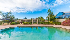 A clear swimming pool with lounge chairs and a covered seating area, surrounded by greenery and modern buildings. - Harrington River Lodge (Harrington)