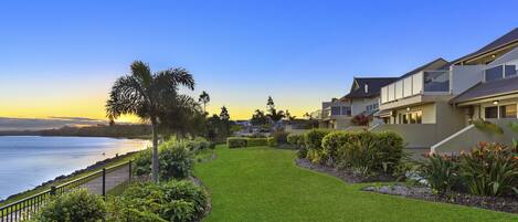 A coastal residential area with a well-maintained lawn, a palm tree, and houses with balconies overlooking the sea.