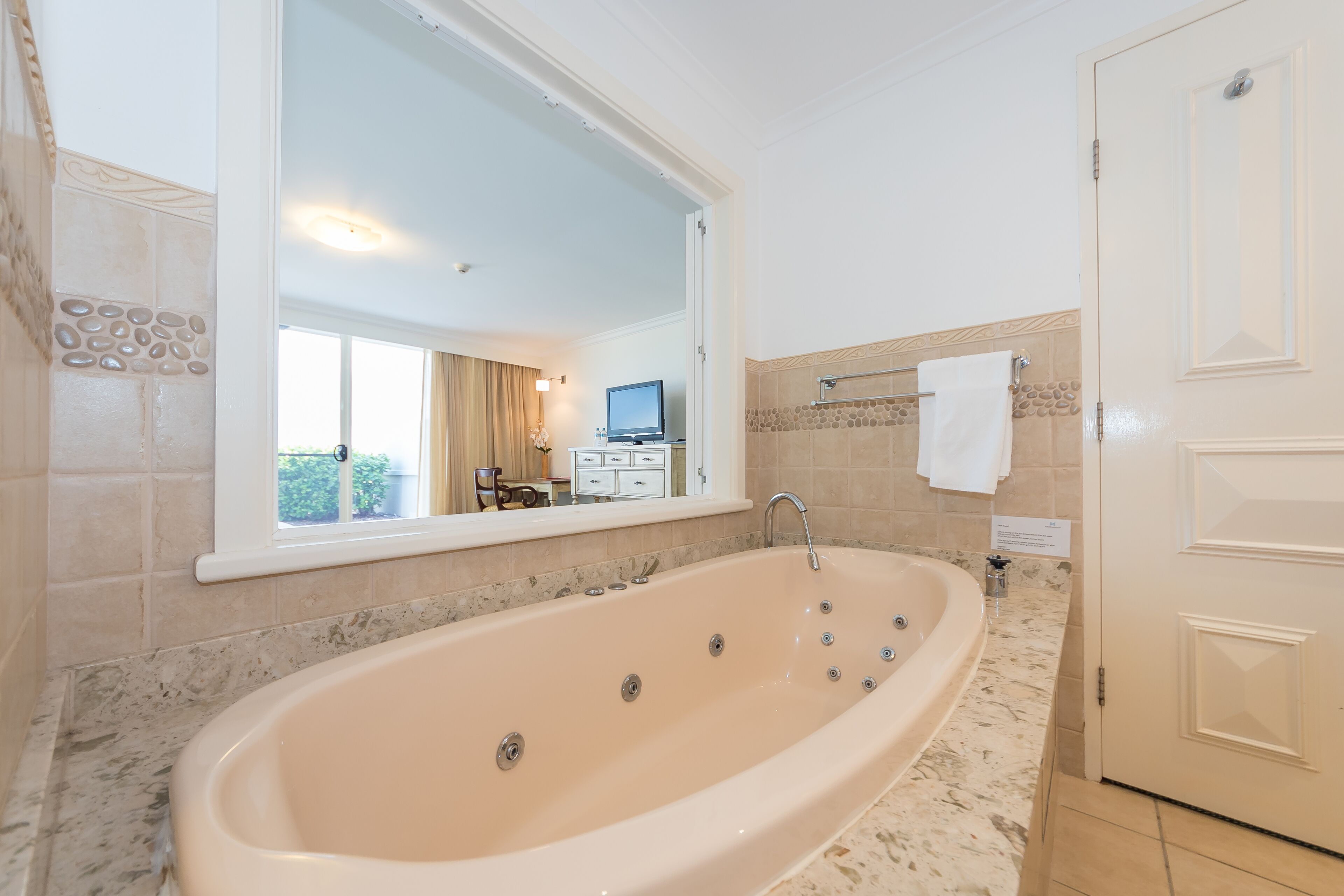 A bathroom with a large bathtub, a mirror, a television, and a window with curtains.