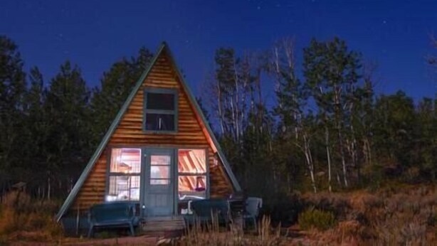 Una cabaña de madera con techo triangular, una pequeña ventana y una puerta. Hay dos sillas afuera de la cabaña.