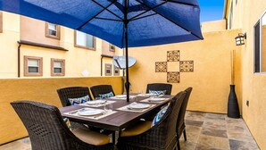 Zona pranzo con tavolo apparecchiato per quattro, circondato da sedie in vimini, sotto un ombrellone blu.