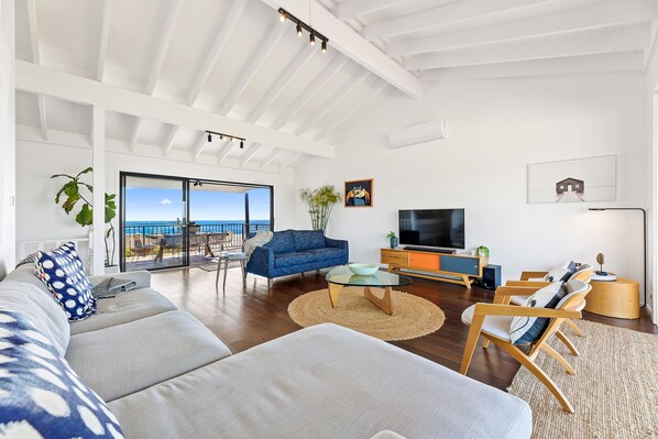 Ein modernes Wohnzimmer mit einer großen Couch, einem Couchtisch und einem Fernseher. Ein Balkon bietet Blick auf das Meer.