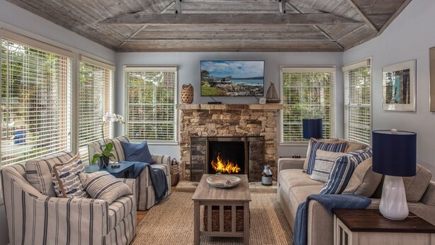 A cozy living room with a stone fireplace, striped sofas, and large windows with blinds.